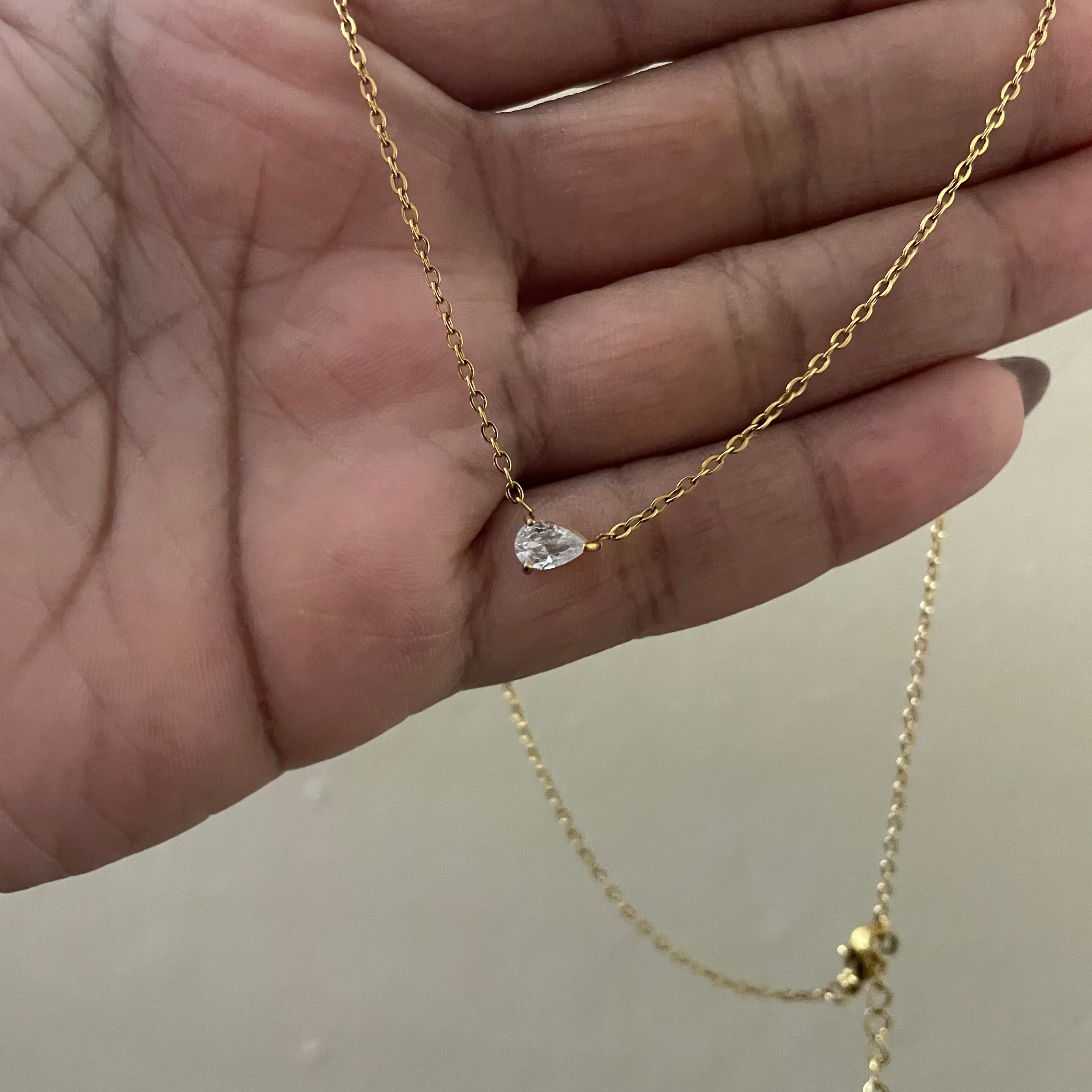 Gold necklace with a teardrop-shaped pendant held by a hand against a neutral background